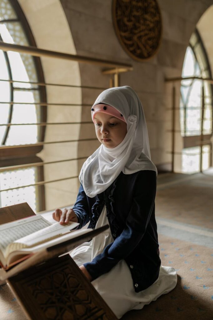 A young girl in a hijab reads the Quran inside a mosque, highlighting Muslim culture and spirituality.